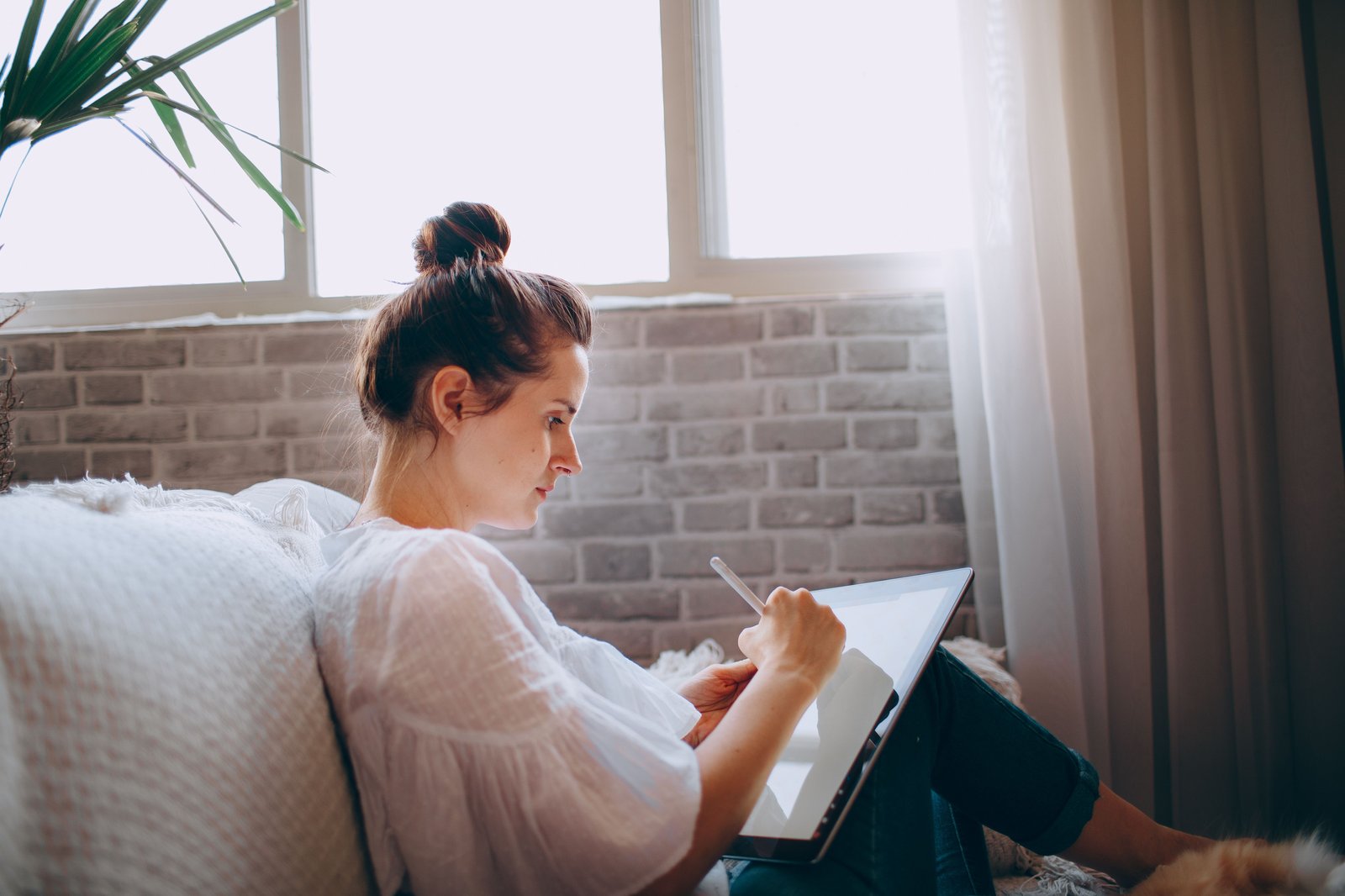 Woman sitting by a sunlit window, quietly writing on a tablet in a cozy living room
